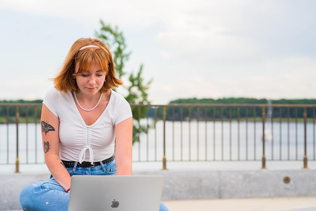 A student sites in front of the water on a bench working on a laptop